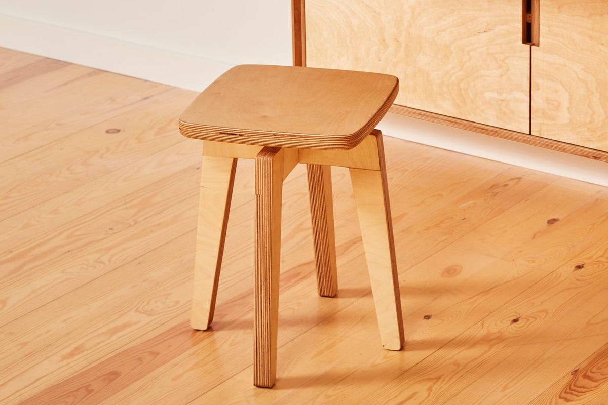 A light natural wood folding desk with a matching wooden stool, set against a minimalist beige wall on a light wood floor. The desk features a drop-down tabletop, two cabinet doors, and a small dark vase with dried stems on top.